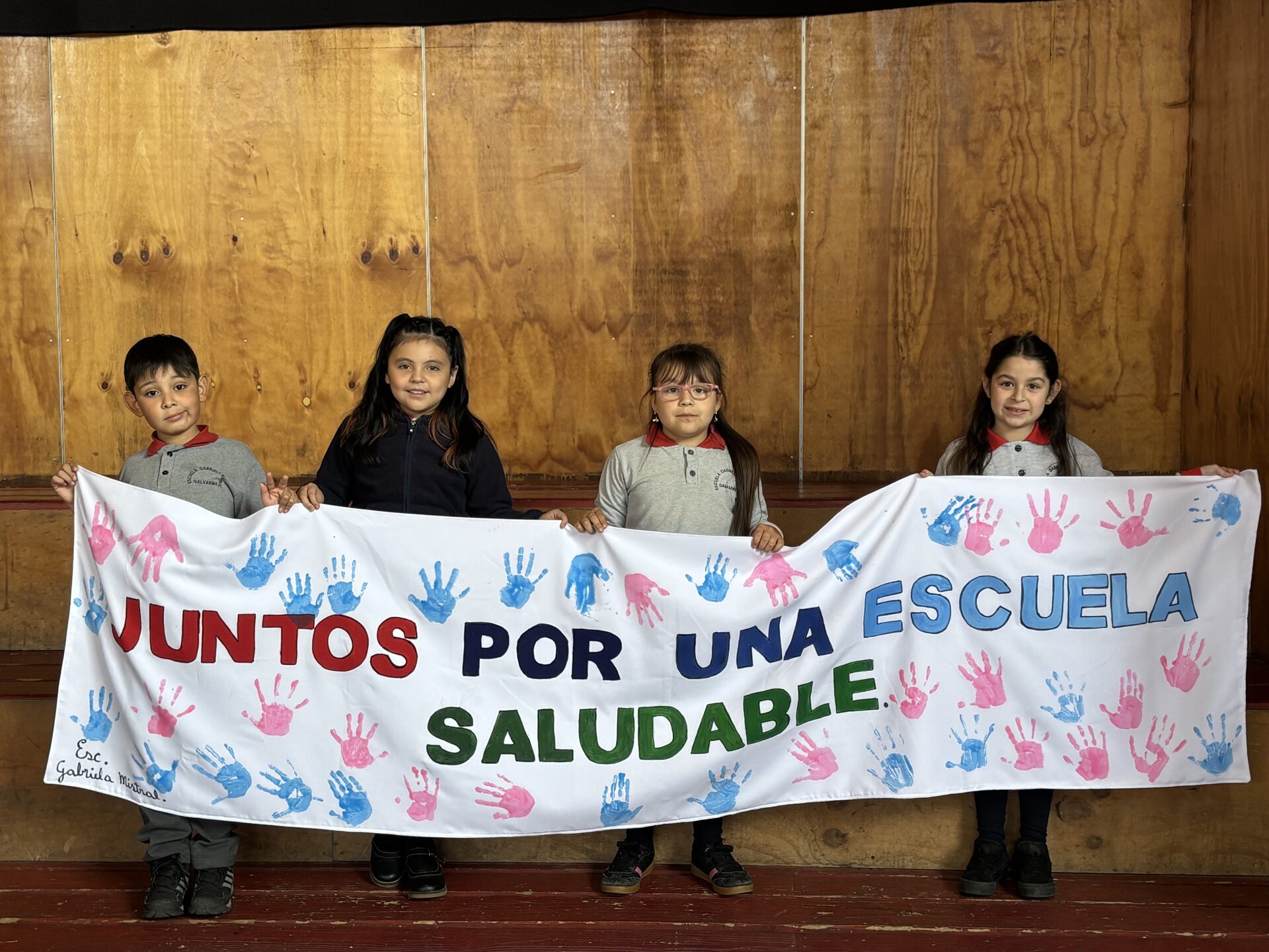 Cuatro estudiantes de párvulos sosteniendo un lienzo "Juntos por una escuela saludable" con huellas de manos pintadas