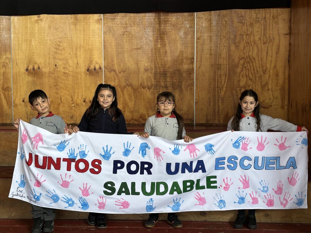 Niñas y niños sonriendo mientras sostienen un lienzo con la consigna "Juntos por una escuela saludable"